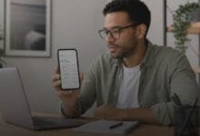 Pessoa consultando saldo disponível no celular enquanto organiza o orçamento financeiro em uma mesa com notebook e caderno.